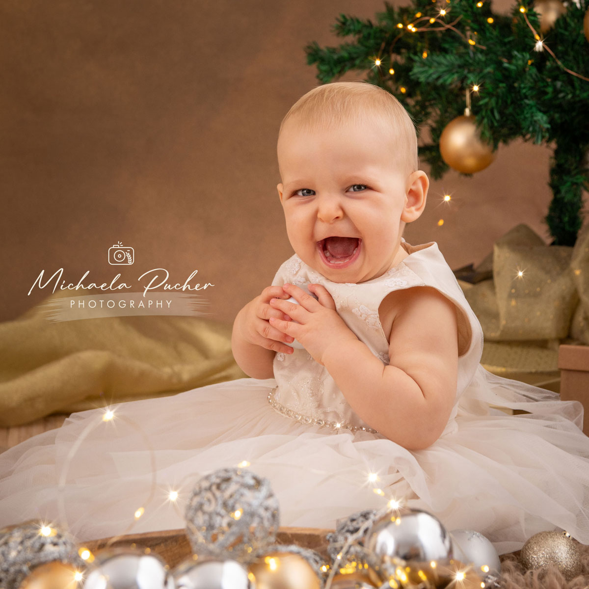 Lachendes Baby im festlichen weißen Kleid sitzt vor einem Weihnachtsbaum, umgeben von glitzernden Christbaumkugeln und warmen Lichterfunken.